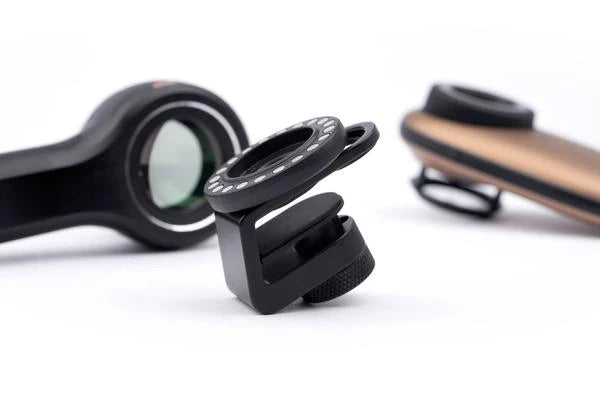 The image shows a black DermLite MagnetiConnect Clamp Adapter (MCC) with a magnetic array, displayed alongside a smartphone camera and a tablet, illustrating its use.