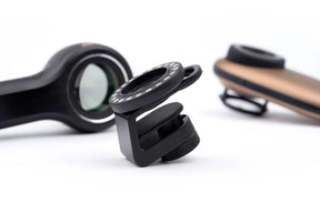 The image shows a black DermLite MagnetiConnect Clamp Adapter (MCC) with a magnetic array, displayed alongside a smartphone camera and a tablet, illustrating its use.