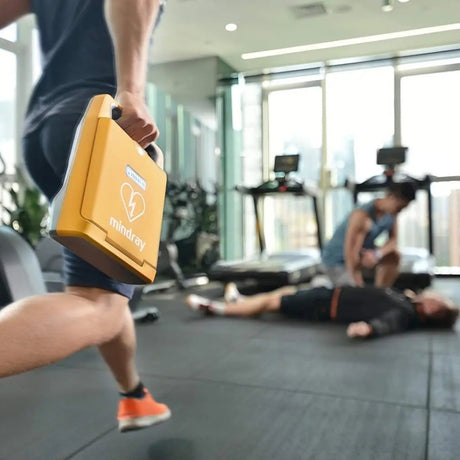 Person holding a yellow AED (Automated External Defibrillator) in a gym setting with another person lying on the floor.